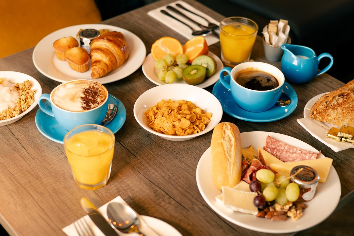 Overhead view of a hotel breakfast spread with a cappuccino and black coffee in blue cups, orange juice, fresh fruit, croissant and muffins, a bowl of cornflakes, yoghurt and granola, and a charcuterie plate with cheese, ham, salami, grapes, nuts and bread.